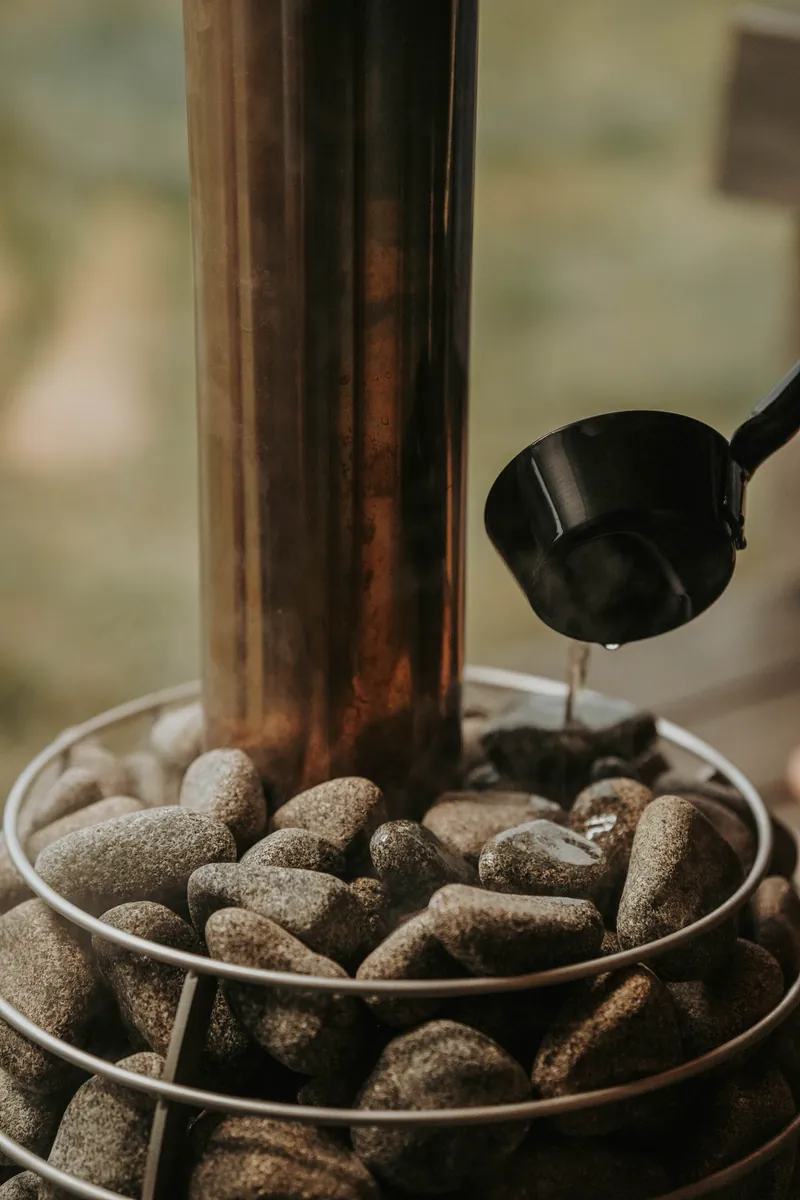 Wooden ladle pouring water onto dark sauna stones with löyly steam rising