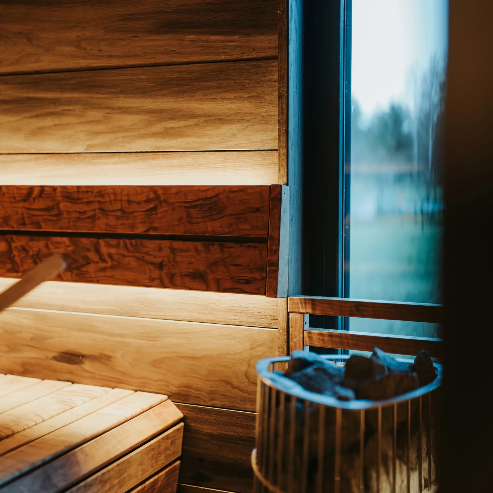 Honey-toned cedar paneling and a slatted bench glow under a warm amber LED inside a sauna, with a stone-filled heater in the foreground and cool daylight through a side window